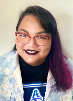 Woman with purple hair wearing red rimmed glasses and whitewashed jean jacket over navy t-shirt