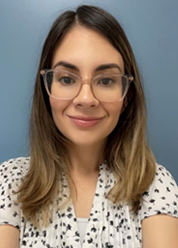 Woman with medium length straight brown hair wearing clear pink rimmed glasses and a white blouse with black polka dots