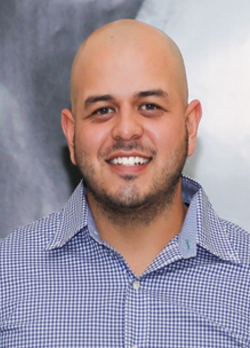 Man with shaved head and stubble beard wearing a blue and white checkered button up shirt