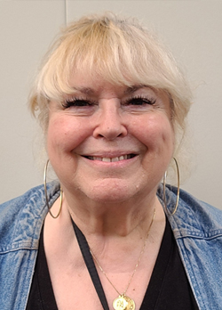 Woman with light blonde hair wearing a denim jacket over black shirt and gold necklace